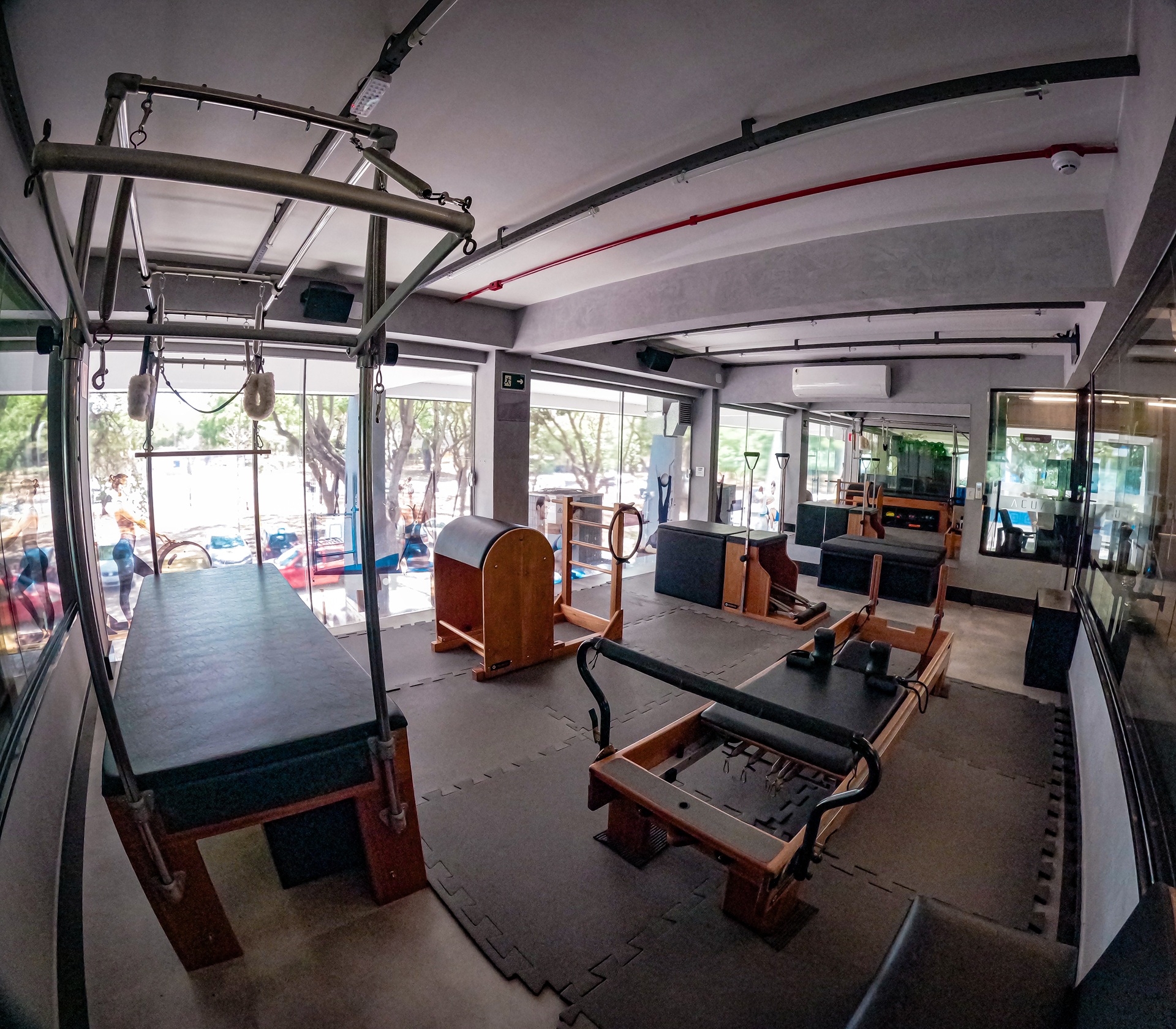 Aparelhos de Pilates alinhados em sala envidraçada com vista para a rua arborizada